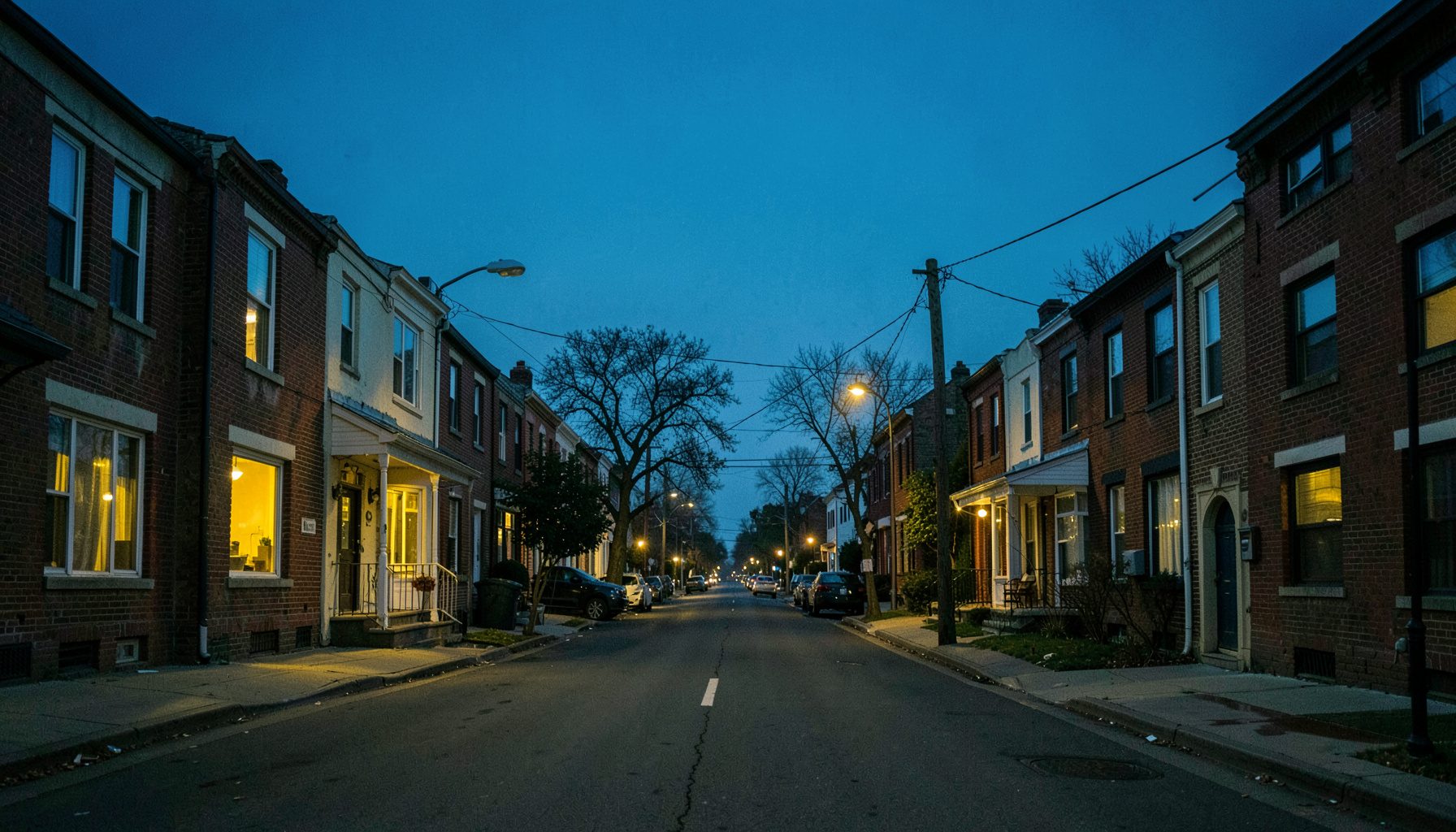 A wide documentary-style photograph of a Toronto neighborhood street at dusk, with rows of modest brick houses and apartment buildings under a deep blue sky. Warm yellow light spills from windows, and the street is quiet. The mood is calm but somber, emphasizing community and place.