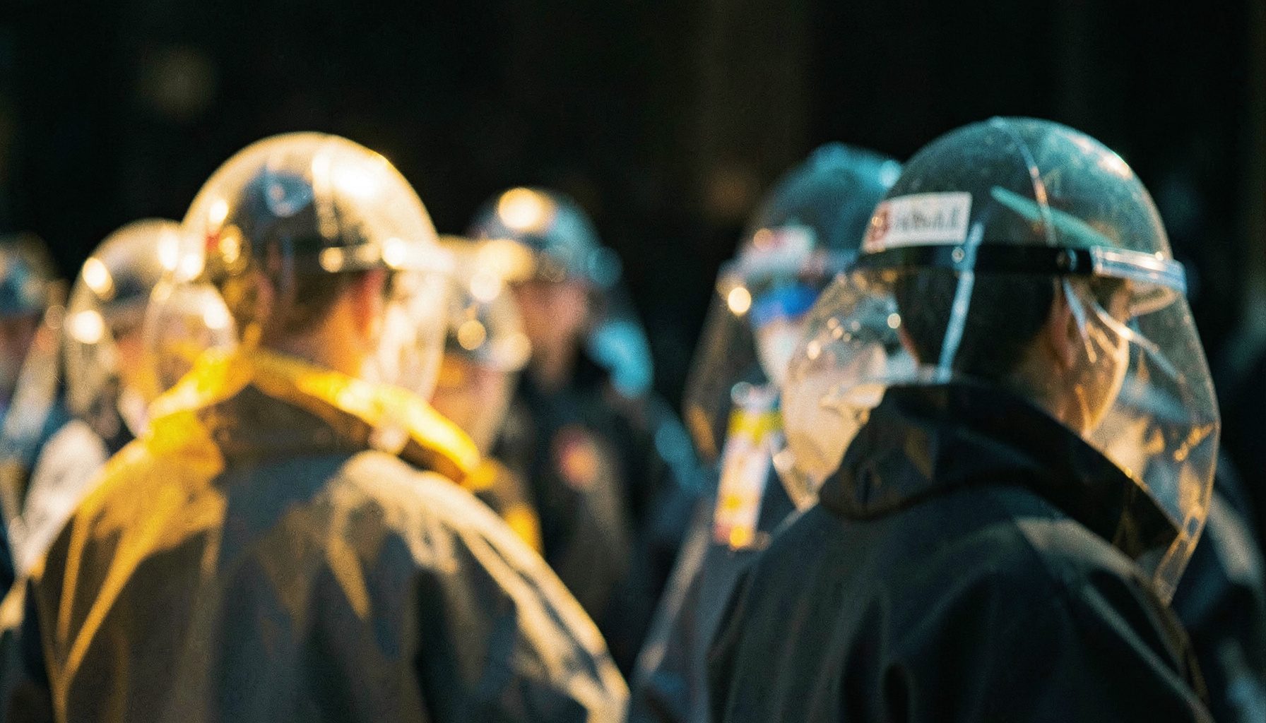 A documentary photograph of essential workers in protective gear, seen from behind in soft focus, with warm amber and cyan lighting against a dark background.